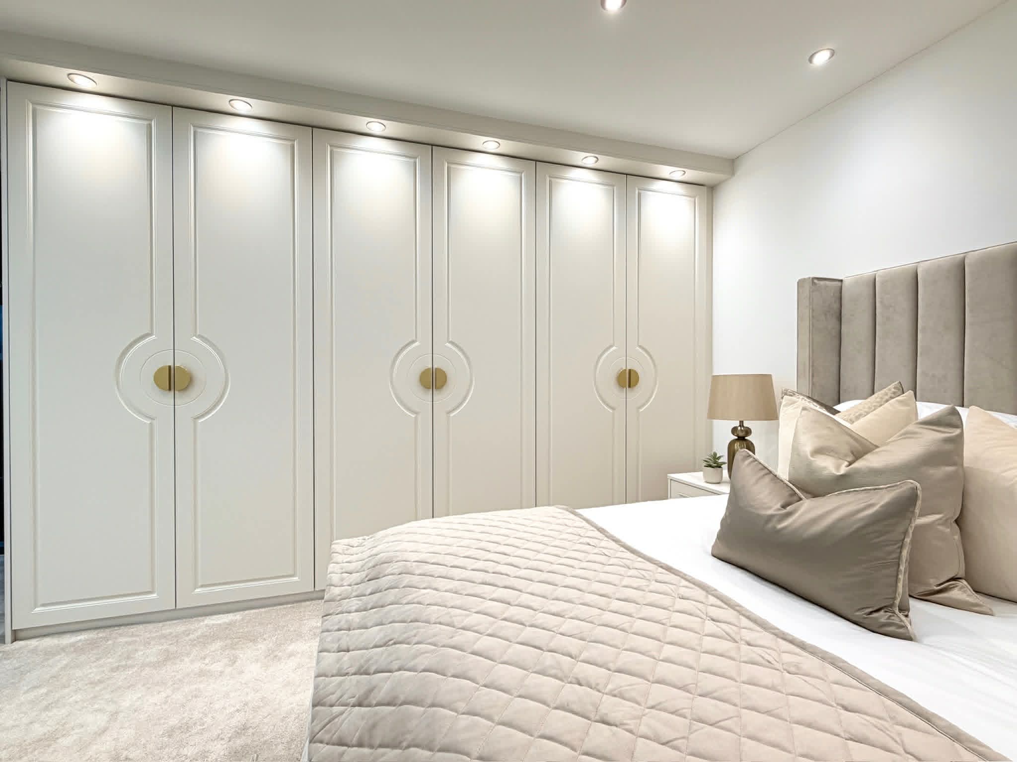 A wall of off-white fitted wardrobes featuring a unique circular carved detail around central gold half-moon handles, illuminated by integrated ceiling spotlights.
